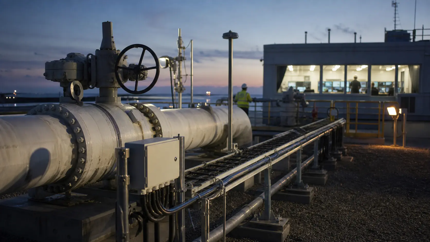 Industrial pipeline control equipment at dusk with data conduits and distant operators, illustrating operating-record boundaries behind pipeline infrastructure.