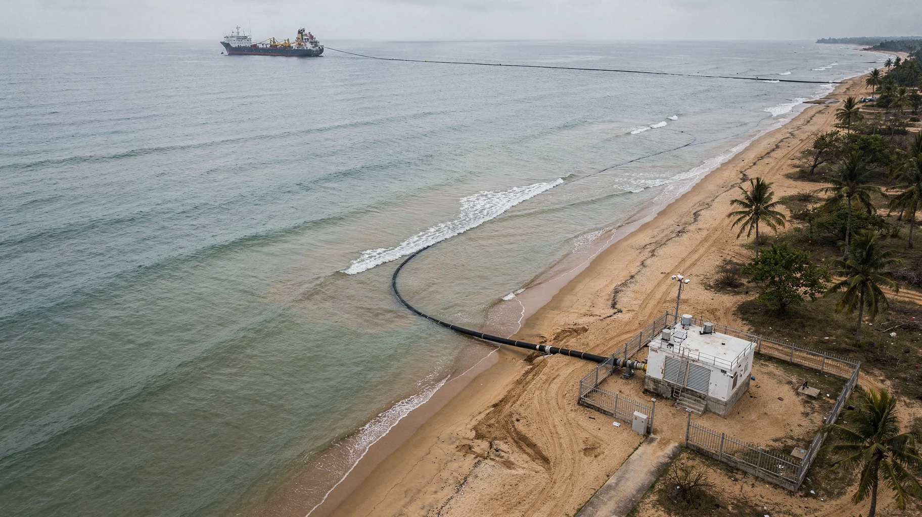 Aerial view of a tropical South Asian coastline where a submarine fibre-optic cable comes ashore at a low concrete landing station with a fenced compound and equipment shelter, palm trees and a wide sandy beach, the black cable visible in the shallows and a cable-laying ship offshore.
