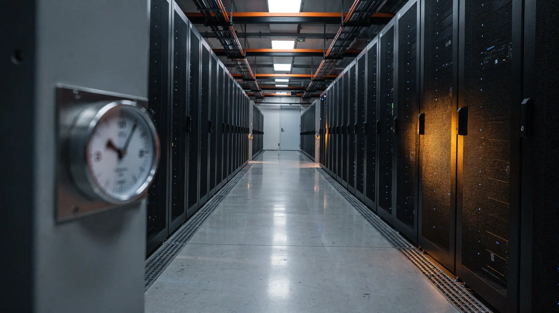 A long cold aisle of black GPU server racks recedes toward a vanishing point under cool industrial lighting, with a small out-of-focus steel dial gauge mounted in the foreground.
