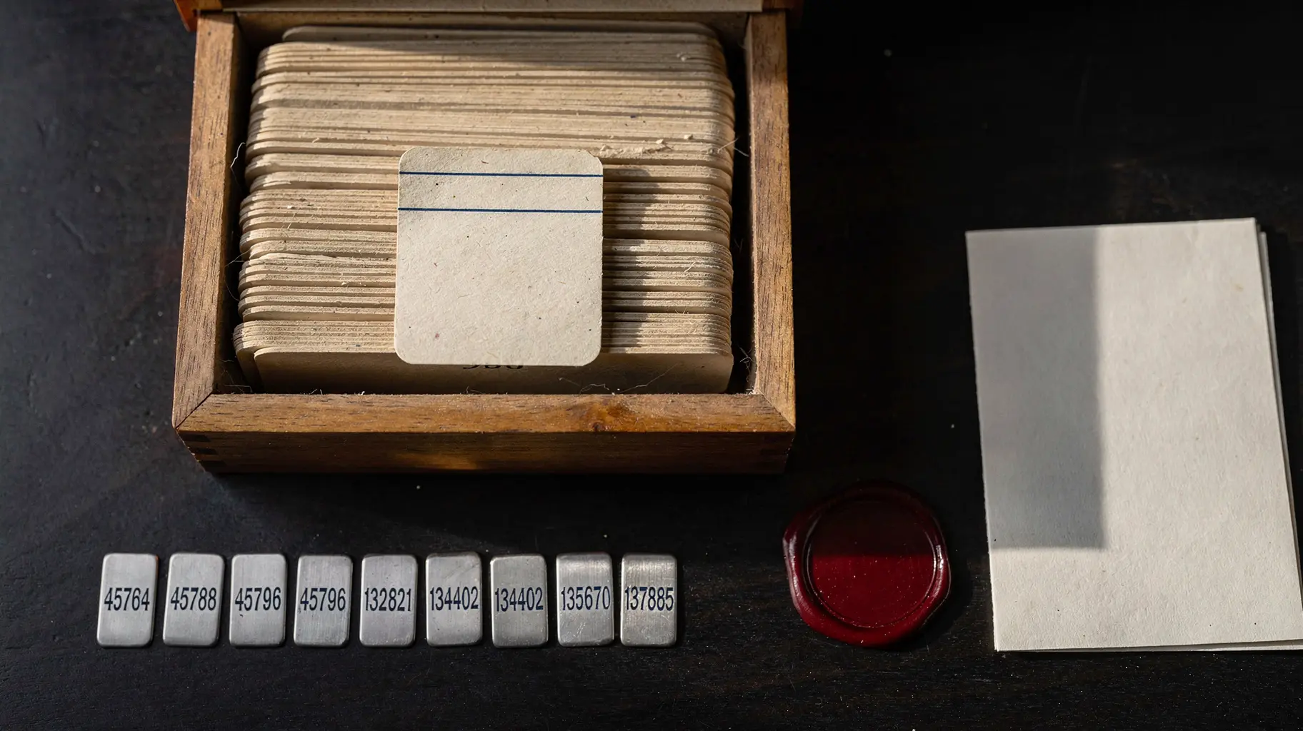 Overhead still life on a dark archival desk: an open card index of aged buff registry cards, one card drawn forward with a single ruled contact line inked in dark blue, seven small numbered metal tabs beside it, and to the right a blank wax seal disc with no impression plus a folded sheet of unmarked paper.