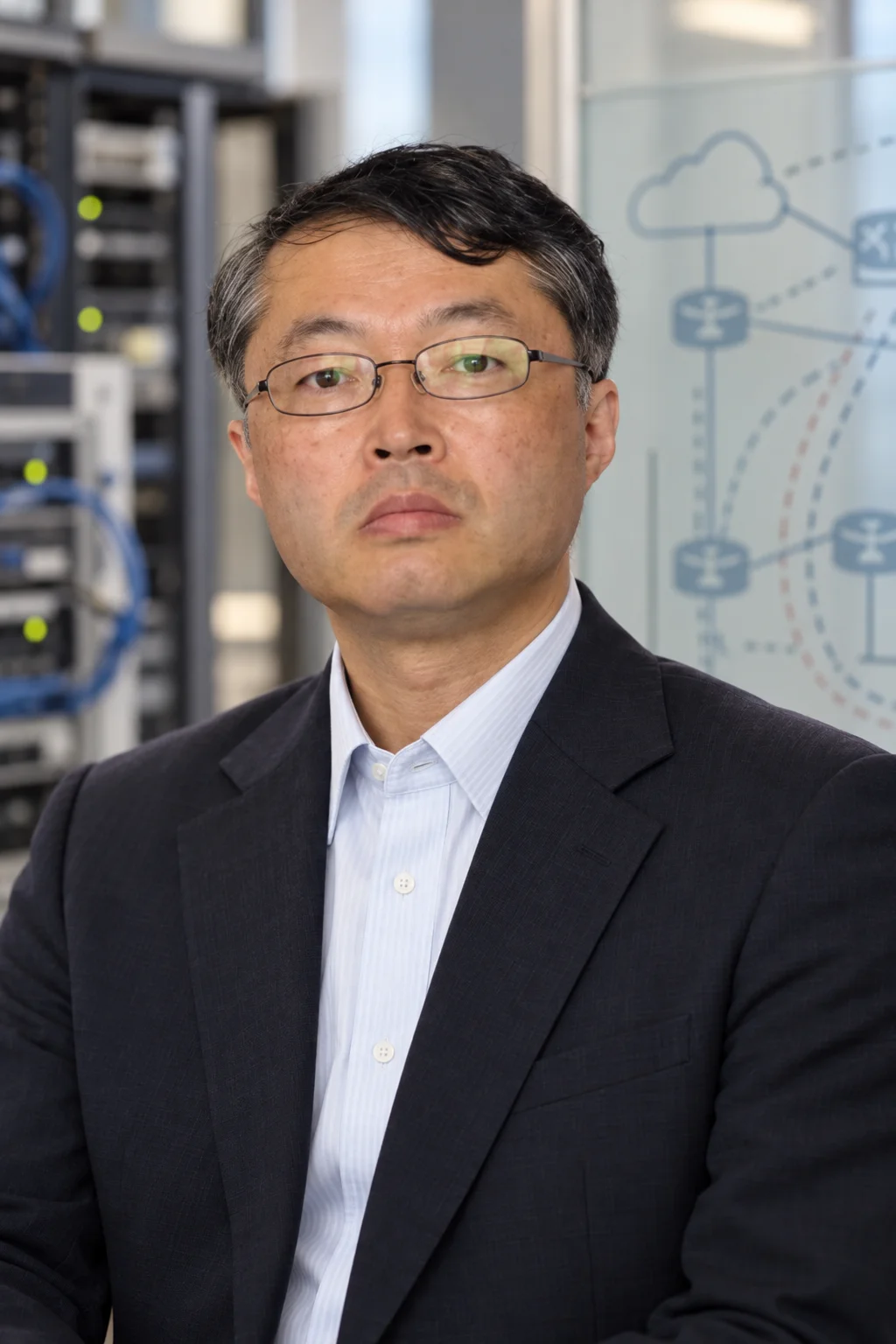 Tomohiro Fujisaki in a head-and-shoulders editorial portrait with a softly focused network engineering workspace behind him.