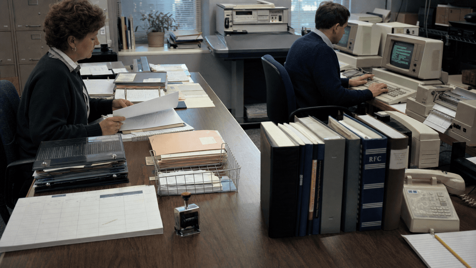 Two 1980s network information centre staff review administrative files beside period terminals, printers, a telephone and technical binders.