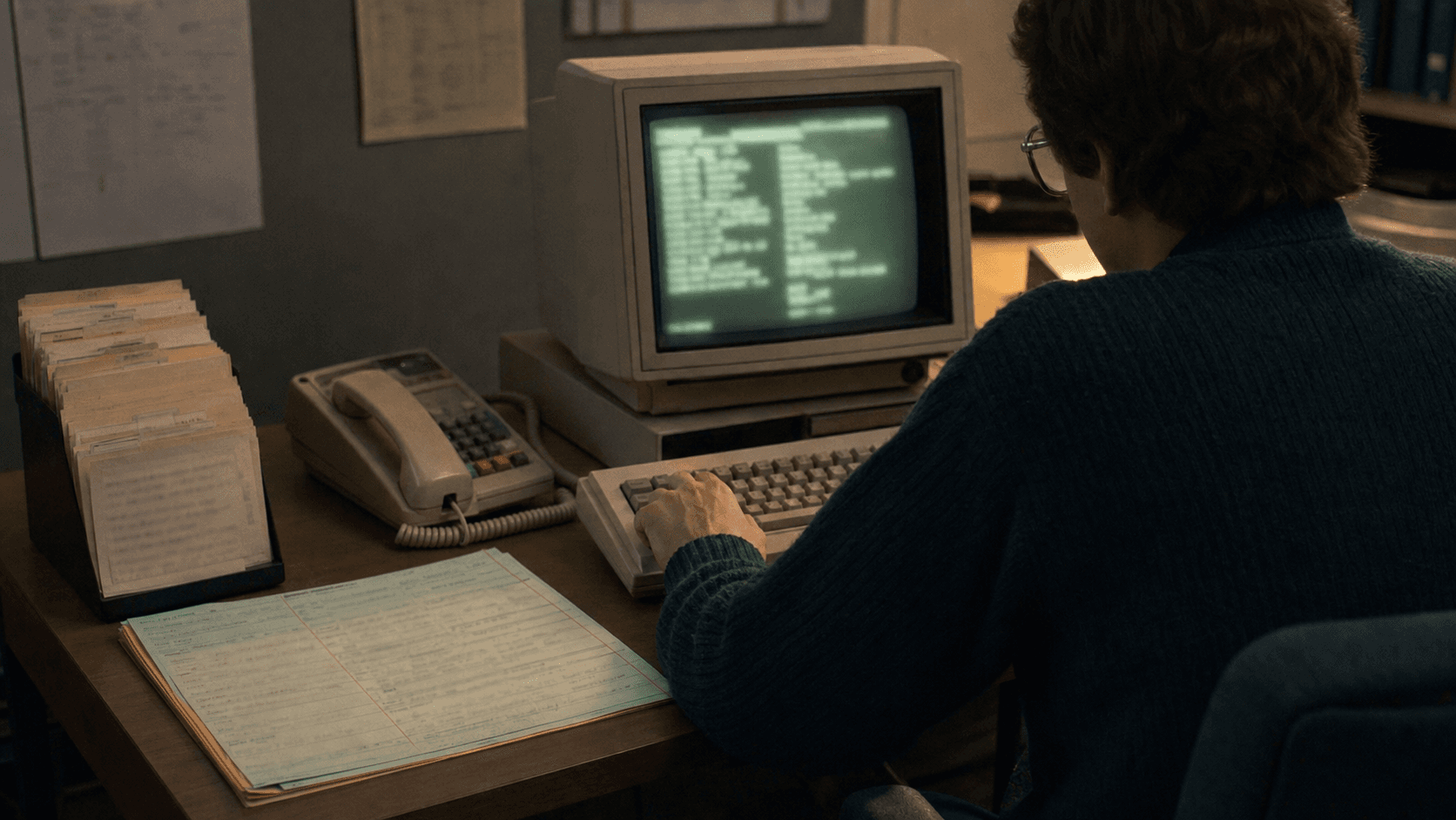 A late-1980s network operator searches a blurred contact directory on a monochrome terminal beside a corded telephone, paper contact file and pending correction form.