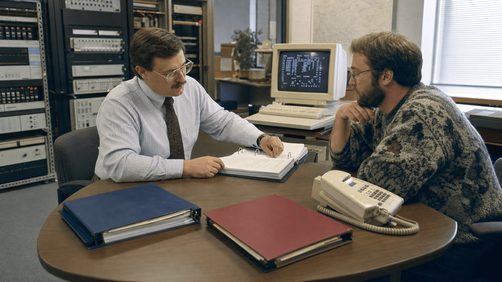 An early-1990s network administrator and engineer review a policy binder and two separate connection folders beside a period terminal and corded telephone.