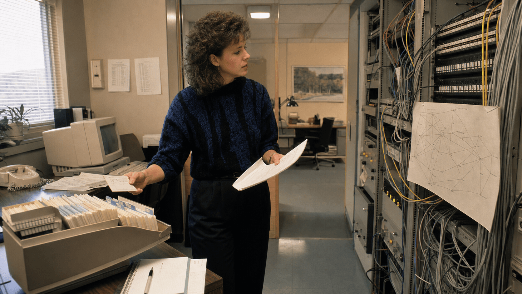 A late-1980s network engineer compares registration cards with a paper topology beside a period terminal, telephone and regional-connectivity equipment rack.