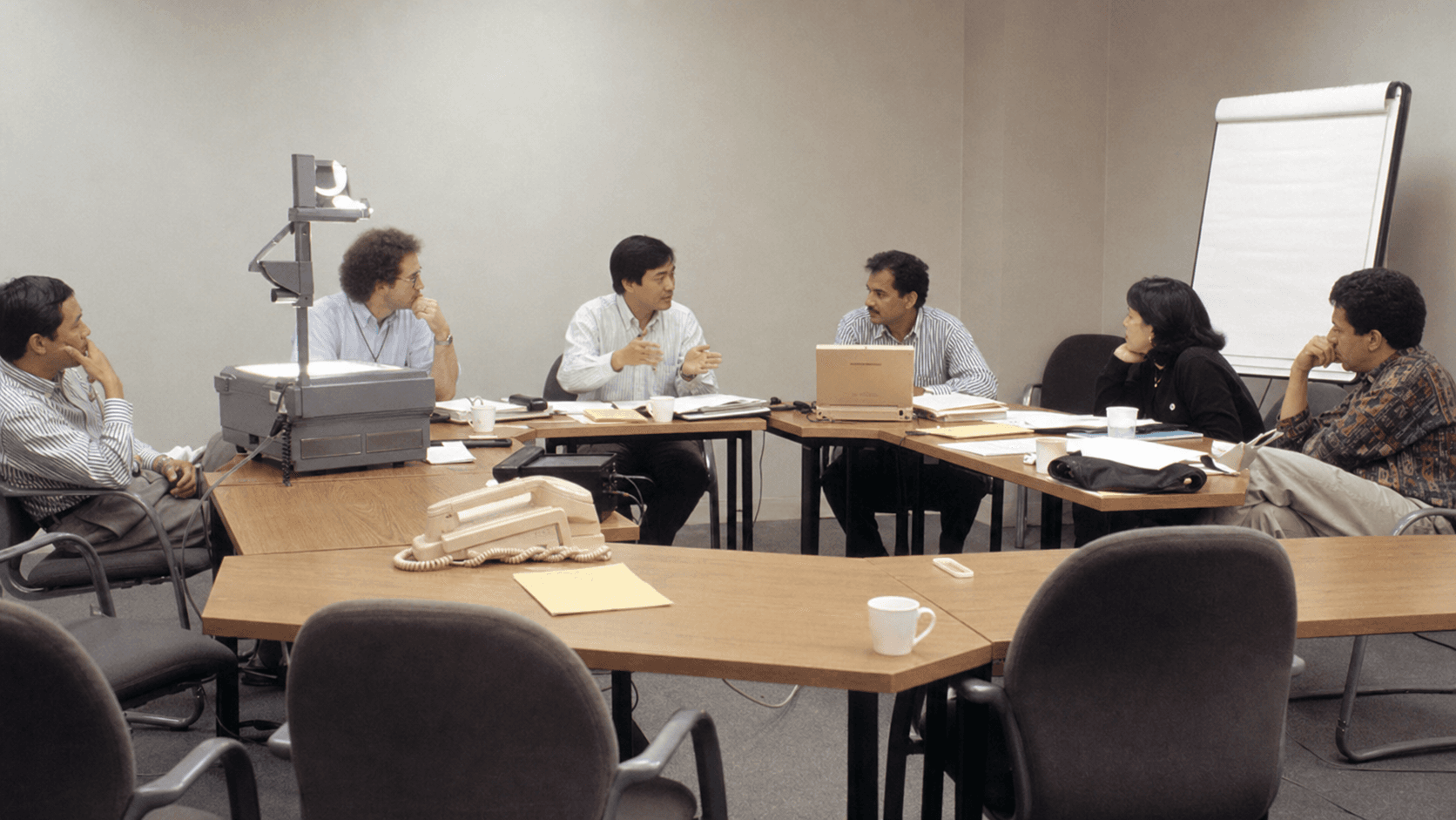 A realistic mid-1990s regional networking meeting with a small multinational group, mismatched tables, an overhead projector and empty chairs.