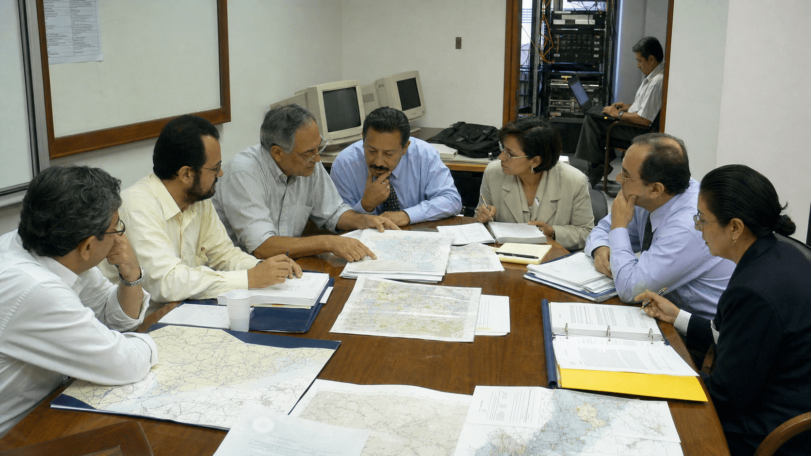 A late-1990s Latin American regional planning meeting with delegates comparing transition folders beside borrowed network equipment.