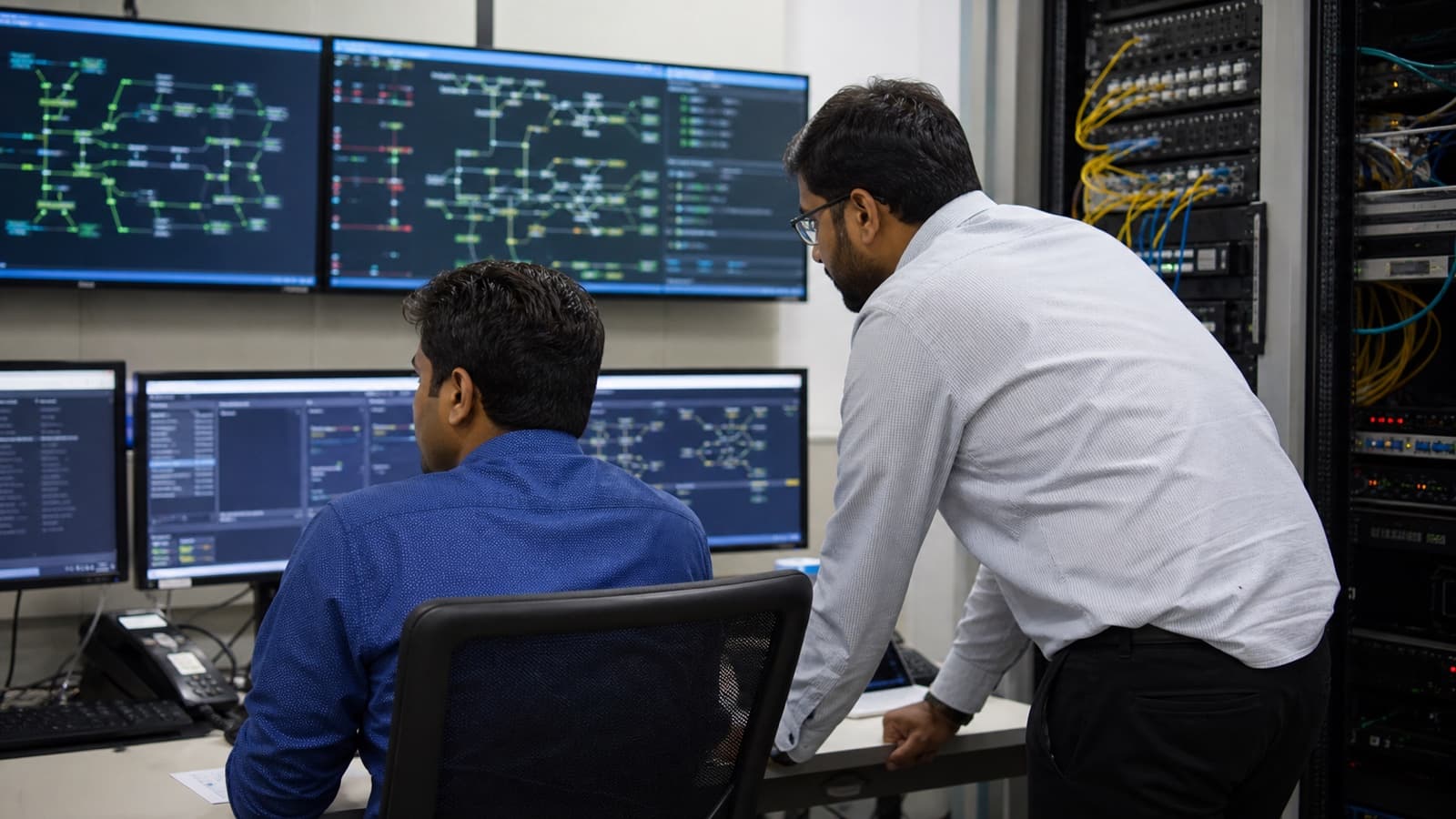 Two network operations engineers reviewing generic network monitoring displays beside telecom equipment racks.