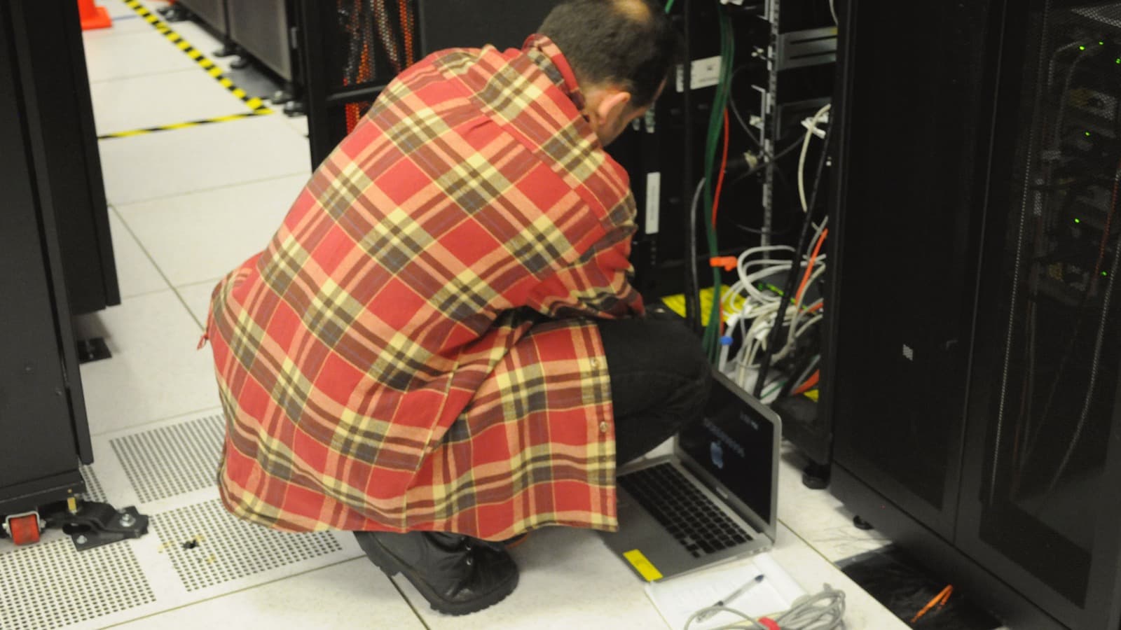 A technician uses a laptop beside server racks in a computing facility