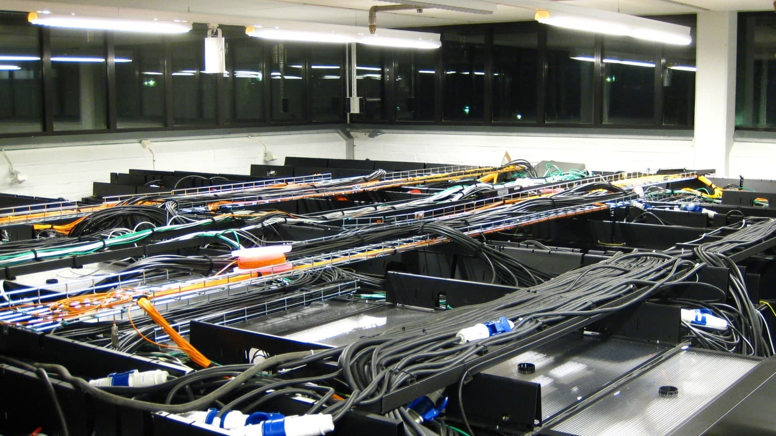 A server room with rows of network equipment, power cabling, and orange and green fiber connections.